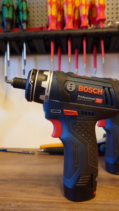 En person håller en Bosch batteridriven skruvdragare med en grön indikatorlampa tänd; verktyg i bakgrunden.