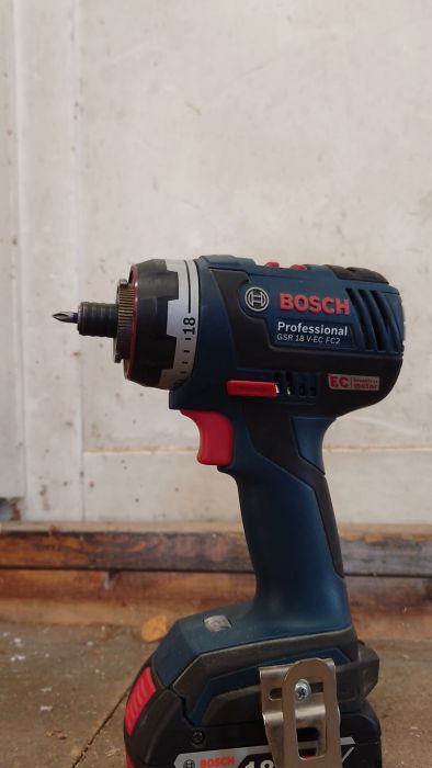 En hand håller en Bosch professionell borr/skruvdragare mot en vit vägg med ett träelement och betonggolv i bakgrunden.