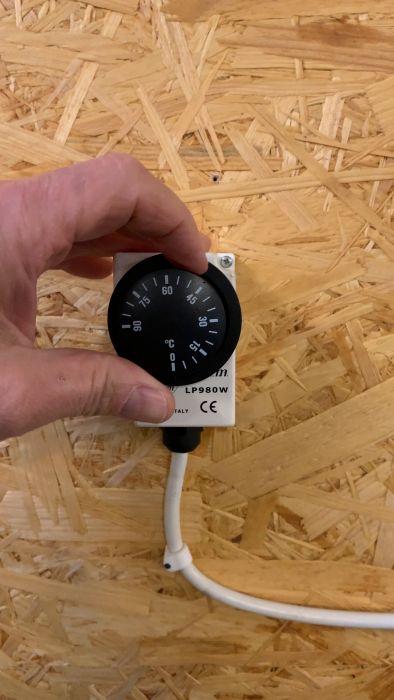 En hand håller en timerbrytare med märkningen "LP980W", monterad på en spånskivevägg med synlig vit kabel.