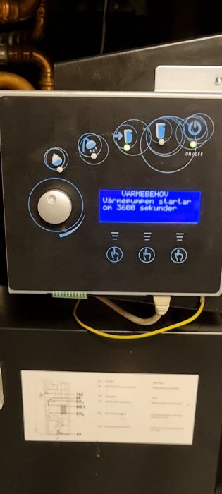 Kontrollpanel för en värmepump med ett blått displaymeddelande, ikoner och knappar och en installationsguide nedanför. Displayen visar en timer.