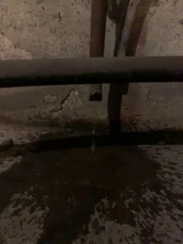 En rörledning läcker vatten på en våt betongyta inne i en mörk, till synes gammal och ounderhållen byggnad.