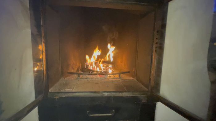 En eldstad med brinnande ved, synlig genom en öppen lucka, med lågor som spricker och fladdrar, och glödande kolet nedanför.