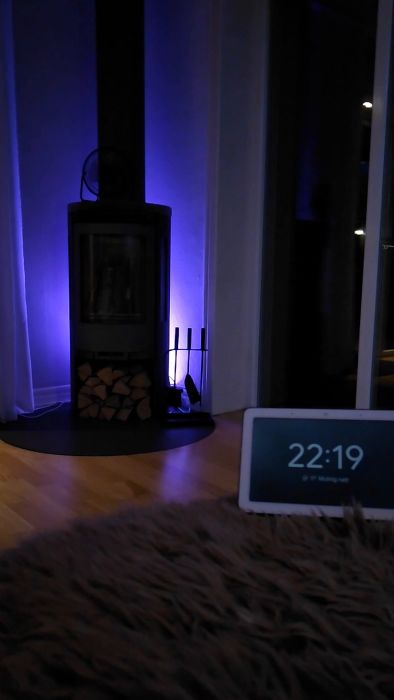 Stämningsfullt vardagsrum med lila ljus, braskamin, ved, klocka som visar 22:19, matta i förgrunden.