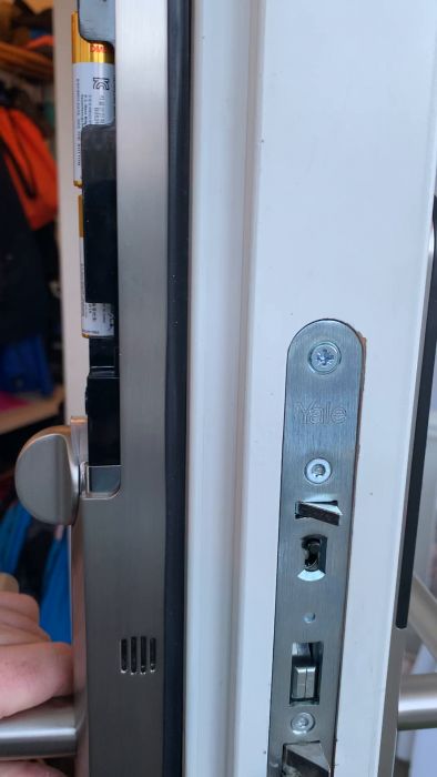 Ett närbildsfoto av en dörrstängare och AA-batteri för att demonstrera storlek. Metall, vit dörrkarm och färgglada bakgrundsobjekt syns.