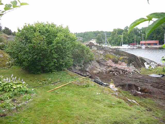 Ett jordområde nära vatten med växtlighet. Synlig erosion eller jordskred med lera och vatten som strömmar ner mot floden eller sjön. Grönområde i förgrunden.