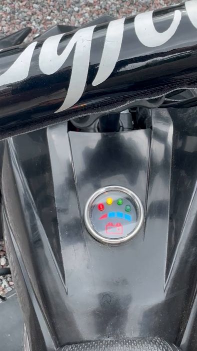 En motorcykelkåpa med dekorativa vita streck och en cirkulär statusindikator visande flera färgade lampor och symboler mot en grusig bakgrund.