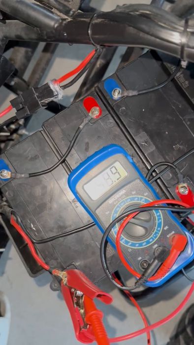 Digital multimeter visar 49.9 volt, ansluten till ett svart batteri med röda och svarta testledningar.