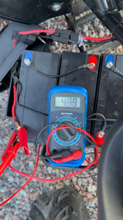 Film på ett multimeter som visar 26.9 volt, anslutet till ett batteri, suddig bakgrund av vad som kan vara fordonets delar.