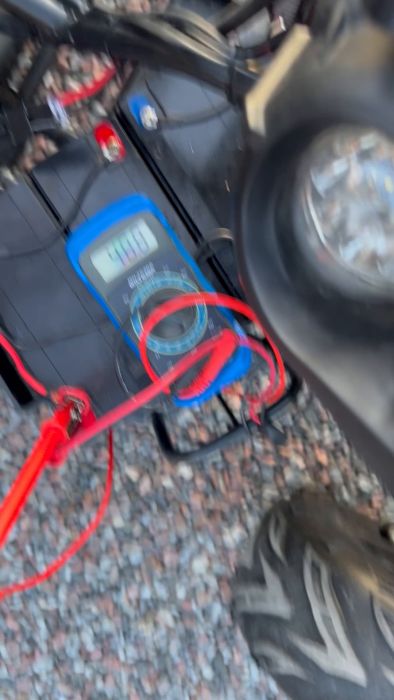 Multimeter kopplat till ett fordonbatteri, visar spänning på 44,7 volt, troligen underhåll eller felsökning av elsystemet på en elektrisk skoter eller motorcykel.