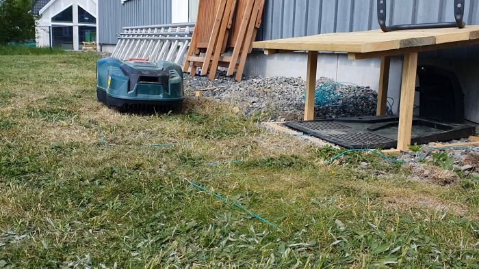 En robotgräsklippare på en gräsmatta nära ett hus, trädgårdsmöbler och material i bakgrunden.