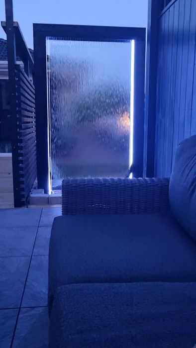 Utomhusplats under kvällstid, vattenfunktion med bakbelyst glas, modern soffa, lugn atmosfär, blåtonade färger.