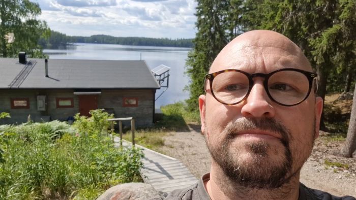 Man tar selfie framför sjöutsikt med grönska och en byggnad vid strandkanten. Soligt och klart väder.