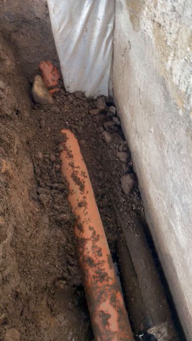 En hand pekar på ett orange rör bredvid ett grått rör i en grävd schakt i marken. Det ser ut som ett byggarbete.