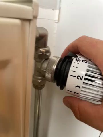 En hand som justerar temperaturinställningen på en vit termostat på en radiator, bredvid en vit vägg.