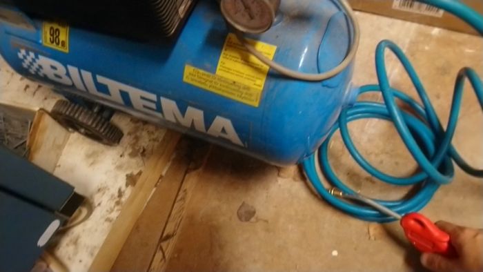 Blå luftkompressor av märket Biltema med tryckmätare och slangar, ser använd ut i ett verkstadsliknande utrymme.