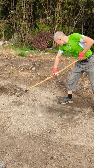 En person i handskar använder en grep med långt skaft för att bearbeta jord i en trädgård eller på en byggarbetsplats.