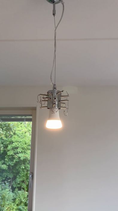 En taklampa hänger med sladdar och clips i rörig formation, med en tänd lampa som visar spektrumfärger, mot en bakgrund av ett fönster och grönt lövverk.