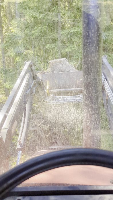 Film inifrån en fordonskabin med utsikt över grävmaskinens arm och skopa i skogsmiljö, smuts på rutan.