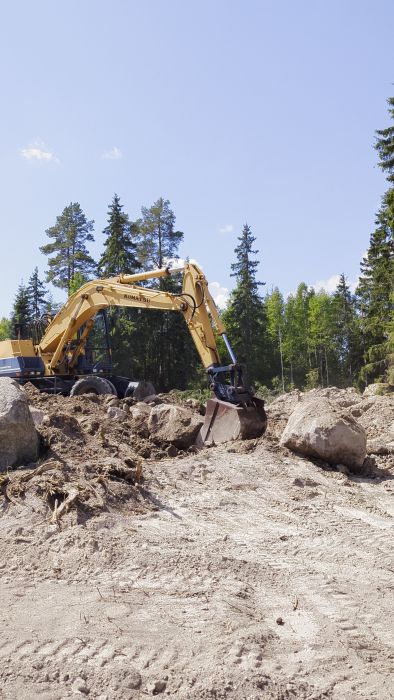 Ett gult grävmaskinsarm med skopa framför en hög med jord och stenar, i ett område som verkar vara under konstruktion eller grävning, med skog i bakgrunden.