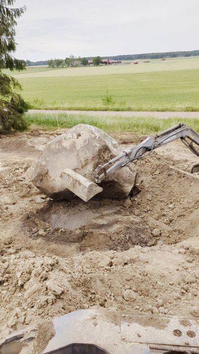 En stor grå klippblock vilar på ojämn, mörk jord. Detaljen och texturerna är tydliga, vilket ger en grov och stenig känsla.