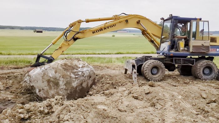 En grävmaskin av märket Komatsu lyfter en stor sten medan en person sitter i förarhytten. De befinner sig på en lerig mark.