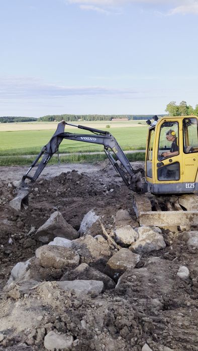 En grävmaskin av märket Volvo arbetar i en jordig terräng med spår av grävarbeten synliga. Ser ut som grävning på en byggplats.