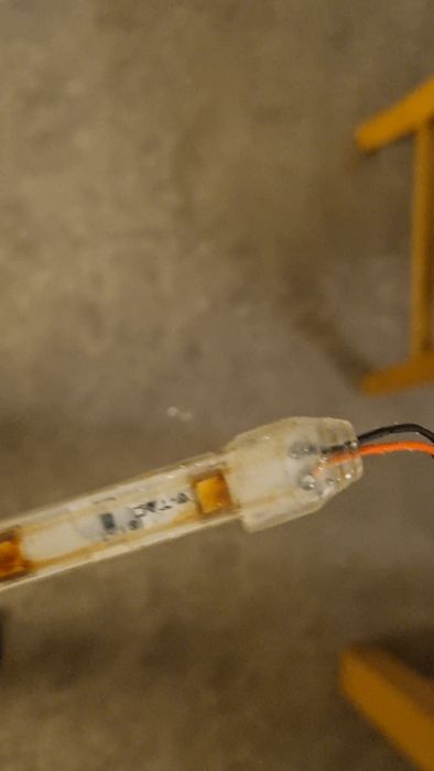 En LED-lampa med synliga kretsar och kablar anslutna, närbild mot en otydlig bakgrund med betong och del av gul stol.