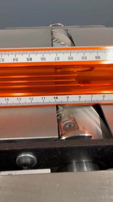 En skruvskruvmejsel mäter en genomskinlig orange linjal placerad på en metallisk yta med synliga skruvar och beslag.