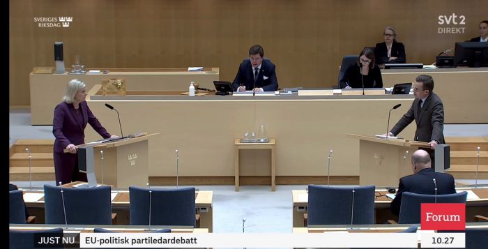 En kvinna i en lila kavaj står vid en talarstol i en parlamentsmiljö. Lyssnare i bakgrunden. Texten indikerar svensk riksdag och TV-sändning.