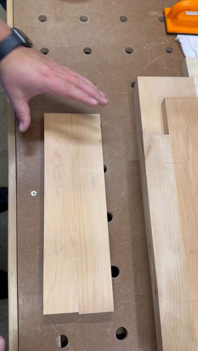 Händer arrangerar träbitar på ett arbetsbord med hål, anteckningar och pilar markerade på träet.