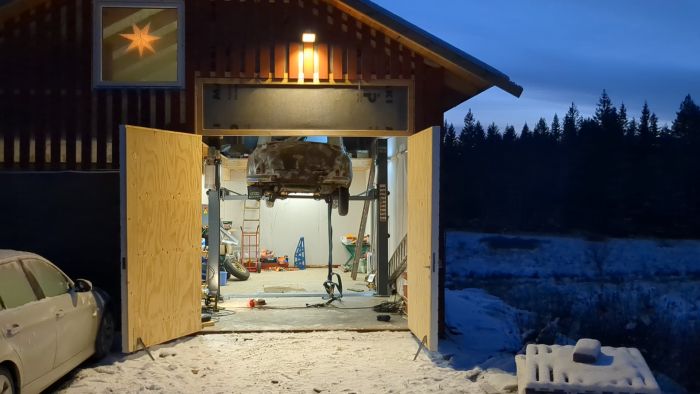 En rostig bil på en lyft i ett rörigt garage. Omgivningen är byggnadsmaterial och verktyg. Filmen är suddig och snett tagen.