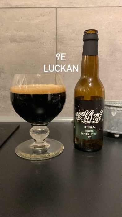 En ölflaska bredvid ett glas mörkt öl på en bänk, med texten "9E LUCKAN" synlig ovanför.