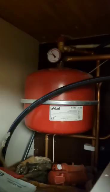Film på en röd expansionskärl för vattensystem med mätare och rör, placerad i ett trångt utrymme med diverse föremål.