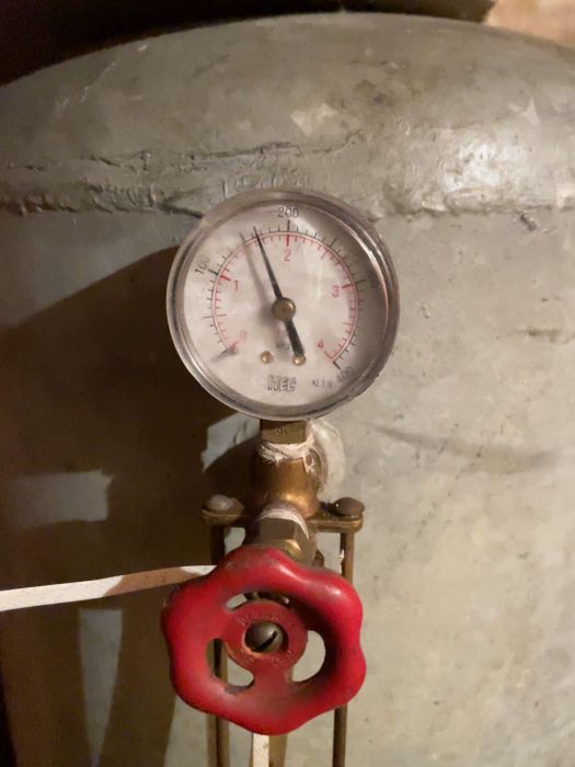 Manometer på en ventil med röd vred för mätning, placerad på en stor metallbehållare, i ett industriellt sammanhang.