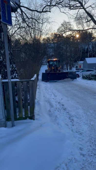 En gatubild med en gul plogbil som röjer snö i skymningen. Det finns snöklädda träd och en staketgrind.