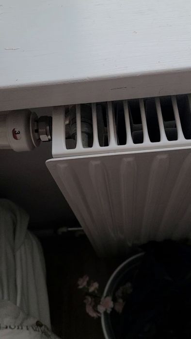 En hand håller i en termostatventil på en radiator under fönsterkarmen. Ventilen är vit och har en röd markör.