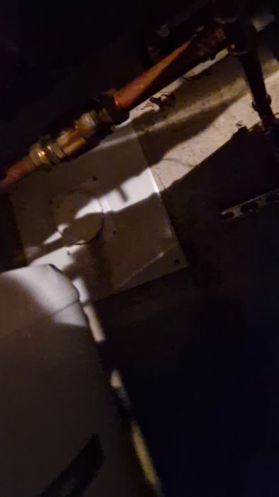 Kopparledning med en backventil monterad på en betongvägg ovanför en vattenberedare, med tecken på vattenläckage nedanför.