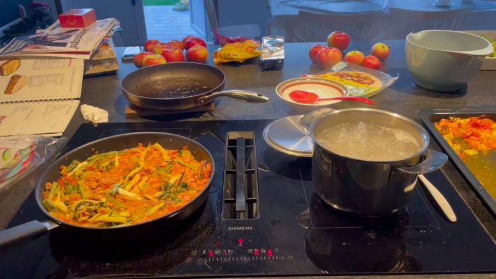 En spis med kokande vatten i en kastrull, en stekpanna med grönsaker och tomatsås, en tom stekpanna, och diverse matförpackningar på köksbänken.