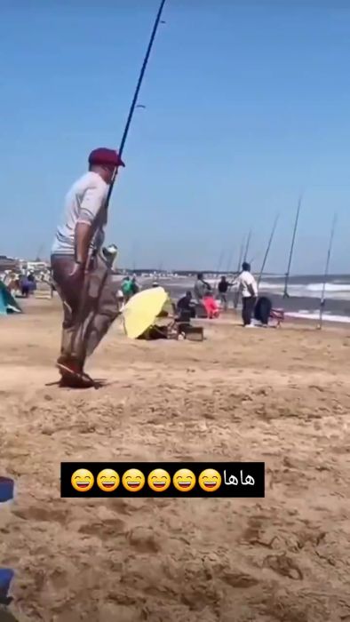 Två personer vid stranden med fiskespön; en sitter, den andra har fallit på alla fyra. Humoristiska emojis och arabisk text nedtill.