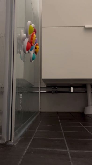 Leksak plan fast i duschvägg, brun kaklad golv, vita väggar och skåp syns i ett badrum.