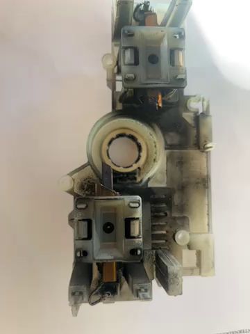 En hand håller en komplex elektronisk komponent mot en vit bakgrund med textdokument. Objektet är troligen en del av en större apparat.