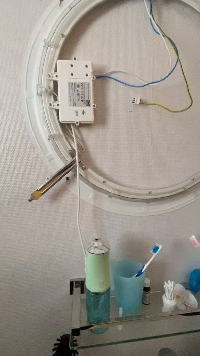 En hand med en strömtestare vid en elektrisk anslutning på en vägglampa, med badrumstillbehör i förgrunden.