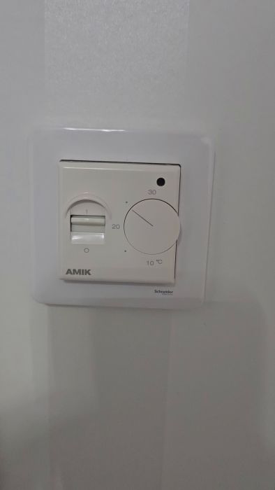 En termostat från Schneider på en vit vägg med en röd lysande indikator, justeringsratt mellan 10 och 30 grader Celsius, märkt 'AMIK'.